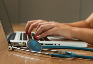 A person typing on a laptop while a stethoscope is placed on the table
